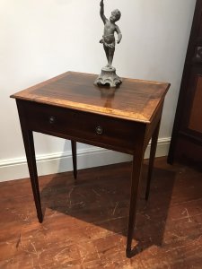 Elegant period antique Sheraton-style side table, circa 1780, featuring a satinwood cross-banded top and a front drawer with original brass handles. One ring handle is missing due to age. Delicate tapered legs are decorated with string inlay, and the drawer has a pine lining. The table has a faded colour and shows signs of wear consistent with age and use. Dimensions: 31.5 inches high × 23 inches wide × 19 inches deep.