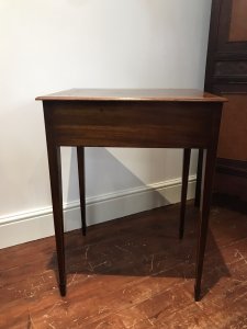 Elegant period antique Sheraton-style side table, circa 1780, featuring a satinwood cross-banded top and a front drawer with original brass handles. One ring handle is missing due to age. Delicate tapered legs are decorated with string inlay, and the drawer has a pine lining. The table has a faded colour and shows signs of wear consistent with age and use. Dimensions: 31.5 inches high × 23 inches wide × 19 inches deep.