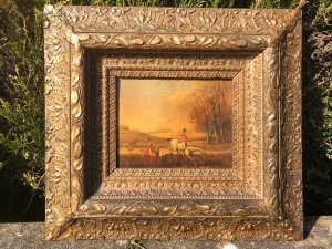 Pair of fine art hunting scene paintings by artist Brain Cooke, circa 1900, executed on oak panels with fine colours and detail. Titled 'The Full Cry' and 'The Chase', the scenes depict mounted hunters navigating water and jumping fences, and red-coated riders galloping with beagles in pursuit. Each painting is presented in an elaborate giltwood frame adorned with fruit, acanthus leaves, roses and orchids. Dimensions not provided.