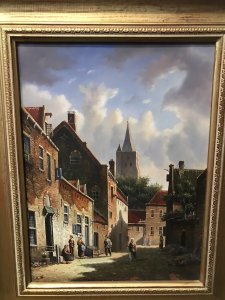 Original Dutch street scene oil painting on board by artist M Ives, presented in a gilt frame. The village view depicts cobbled streets and seven male and female figures in traditional dress, with a spired church in the background beneath a blue sky.