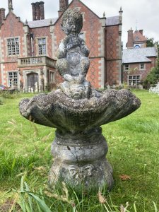 Water Feature Centrepiece Of Faun Boy Sat Upon Scalloped Bowl. Circa - 20th Century Composition- Reconstituted Stone Condition- Weathered Used Pristine Decorative Water Feature Of Scalloped Bowl Set Upon Square Oval Plinth Pediment With Bow And Column Pillared Decoration, With Central Sitting Faun Playing Mouth Organ With Hoove Feet And Bushy Tail. A Hole For Water Hose Through Base Pediment Into Scalloped Bowl. A True Mythological Piece An Asset To Any Garden Decor. Dimensions In Inches Height 30” Width 19” Depth 18” Weight Approx 30Kg Heavy Item
