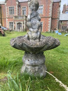 Water Feature Centrepiece Of Faun Boy Sat Upon Scalloped Bowl. Circa - 20th Century Composition- Reconstituted Stone Condition- Weathered Used Pristine Decorative Water Feature Of Scalloped Bowl Set Upon Square Oval Plinth Pediment With Bow And Column Pillared Decoration, With Central Sitting Faun Playing Mouth Organ With Hoove Feet And Bushy Tail. A Hole For Water Hose Through Base Pediment Into Scalloped Bowl. A True Mythological Piece An Asset To Any Garden Decor. Dimensions In Inches Height 30” Width 19” Depth 18” Weight Approx 30Kg Heavy Item