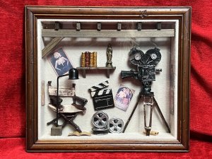Vintage Diorama Of Film Directors Studio With 1920's Icons Circa - Vintage Composition - Mixed Media Paper/Resin/ Glass/ Wood Listing Description Stylish Diorama Of 1920's Hollywood Directors Studio In Room Of Textured Wallpaper With Wood Exposed Beams, Showcasing Large Camera On Tripod Stand. Wall Shelf With Prevalent Oscar Film Award And Books And Walls Dec0orated With Film Advertisements Including 'Grandma's Boy' by Harold Lloyd & 1936 Film 'Desire ' And Hollywood Stars Including Elvis And Marlyn Monroe. The Floor Displayed With Rolled Scripts And Film Reels With Clapboard, A Rustic Foldable Chair Reads 'Director' Next To Ornamental Floor Lamp. Diorama Set Inside Wooden Frame With Wall Hanging Display On Reverse. Fine Diorama For Hollywood Film Enthusiast. Dimensions In Inches Height 13 3/4 X Width15.5" X Depth 3"