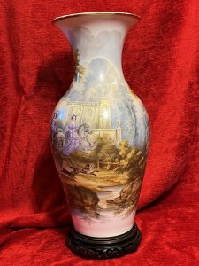 Large Antique Begonia Centrepiece Vase Adorned With Caroline Period Artwork Circa - 19th Century Composition - Porcelain Listing Description Large British Antique 19th Century Decorative Begonia Shaped Vase, Elaborately Decorated With A Romantic Finely Detailed Hand Painted Landscape Scene With Royal Figures Upon Horseback. The Breath-Taking Artwork Depicts A Central Male Figure Sittings Upon Male Bay Horse Wearing Elegant Blouse, Pantaloons, Leather Boots And Hat With Ostrich Plume Posed With Turned Head And Riding Crop Raised In Hand. The Left Gentleman Figure's Galloping Horse Looks Startled, He Wears Identical Outfit In Red And Lilac Hues. (Right Side) Elegant Female In Lavish Lilac Gown With Matching Bonnet With Feather Plume Rides Side Saddle Upon Grey Trotting Horse, Beagles Run Amongst The Horses Legs. A Large Corinthian Columned Palace In The Backdrop. The Landscape Scene Flows Seamlessly Around The Vase Showcasing A Flowing River With British Trees, Rocks And Foliage With Monumental Arched Bridge Crossing The Water. Vase Sits Upon Wooden Plinth. This Impressive Showpiece Vase Is A Fabulous Centrepiece And Talking Point, To Be Displayed Amongst Other Period Treasures. Wonderful Collectors Item. Unsigned Dimensions In Inches Height 19.5" X Width (Widest Point) 9.5" For Further Information Please Call 07376618513 Thank-you