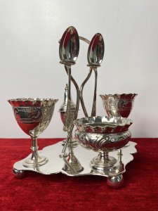 Antique Elegant Egg Cruet Breakfast Set In Christopher Dresser Style. Circa - 1870/75 Composition- EPNS Silver Plate Beautiful Antique Original Breakfast Cruet Set Which Includes Salt Bowl, Pepper Shaker, Two Egg Cups, Two Spoons And Ornate Stand. Delicate Pieces With Scalloped Edges, Ornate Repoussé Work Of Vine Leaves And Scrolls Set Upon Elegant Diamond Clover Shaped Base With Handle, Spoon Holders And Bun Feet. Hallmarked And Stamped. Fabulous Quality Set In Christopher Dresser Aesthetic Movement Style. Dimensions In Inches Height 7” Width 6 1/4” Depth