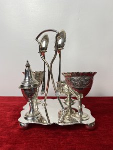 Antique Elegant Egg Cruet Breakfast Set In Christopher Dresser Style. Circa - 1870/75 Composition- EPNS Silver Plate Beautiful Antique Original Breakfast Cruet Set Which Includes Salt Bowl, Pepper Shaker, Two Egg Cups, Two Spoons And Ornate Stand. Delicate Pieces With Scalloped Edges, Ornate Repoussé Work Of Vine Leaves And Scrolls Set Upon Elegant Diamond Clover Shaped Base With Handle, Spoon Holders And Bun Feet. Hallmarked And Stamped. Fabulous Quality Set In Christopher Dresser Aesthetic Movement Style. Dimensions In Inches Height 7” Width 6 1/4” Depth