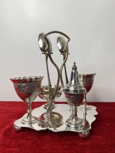 Antique Elegant Egg Cruet Breakfast Set In Christopher Dresser Style. Circa - 1870/75 Composition- EPNS Silver Plate Beautiful Antique Original Breakfast Cruet Set Which Includes Salt Bowl, Pepper Shaker, Two Egg Cups, Two Spoons And Ornate Stand. Delicate Pieces With Scalloped Edges, Ornate Repoussé Work Of Vine Leaves And Scrolls Set Upon Elegant Diamond Clover Shaped Base With Handle, Spoon Holders And Bun Feet. Hallmarked And Stamped. Fabulous Quality Set In Christopher Dresser Aesthetic Movement Style. Dimensions In Inches Height 7” Width 6 1/4” Depth