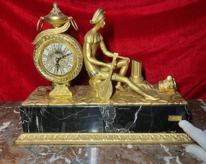 Italian Empire Figural Salon Clock With Posed Court Jester Holding Loot Circa -1950 Composition - Gilt Bronze/ Marble Listing Description A Fine Italian Gilt Bronze Timepiece Showcasing Clock Set Upon Column Mount, With Circular Bezel Face. Enamel Plaque Roman Numeral Dial With Fleur De Lys Decoration And Delicate Folate Hands. Large Ornamental Handled Urn Top Pinnacle And Unusual Sweeping Feather Plume. A Court Jester In Harlequin Costume Sits Against The Clock, Holding Loot Instrument Surrounded By Fallen Column Pillars. Figural Display Is Mounted Upon Black Marble Pediment With Gilt Bronze Frieze. Wonderful Timepiece With Rare Subject. Condition Report Signs Of Tarnishing To Gilt Work Due To Age And Usage. Dimensions In Inches Height 15" X Width 19" X Depth 8" PLEASE NOTE This Is A Very HEAVY ITEM. Due To This Heavy Nature Of This Clock, Delivery Will Be By Hand By Our Curator , Personal Collection By Buyer Or Collection By Specialist Courier Organised By The Buyer. For Further Information Or Enquiries Please Call The Curator On 07707 488566