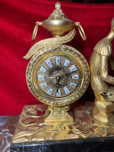 Italian Empire Figural Salon Clock With Posed Court Jester Holding Loot Circa -1950 Composition - Gilt Bronze/ Marble Listing Description A Fine Italian Gilt Bronze Timepiece Showcasing Clock Set Upon Column Mount, With Circular Bezel Face. Enamel Plaque Roman Numeral Dial With Fleur De Lys Decoration And Delicate Folate Hands. Large Ornamental Handled Urn Top Pinnacle And Unusual Sweeping Feather Plume. A Court Jester In Harlequin Costume Sits Against The Clock, Holding Loot Instrument Surrounded By Fallen Column Pillars. Figural Display Is Mounted Upon Black Marble Pediment With Gilt Bronze Frieze. Wonderful Timepiece With Rare Subject. Condition Report Signs Of Tarnishing To Gilt Work Due To Age And Usage. Dimensions In Inches Height 15" X Width 19" X Depth 8" PLEASE NOTE This Is A Very HEAVY ITEM. Due To This Heavy Nature Of This Clock, Delivery Will Be By Hand By Our Curator , Personal Collection By Buyer Or Collection By Specialist Courier Organised By The Buyer. For Further Information Or Enquiries Please Call The Curator On 07707 488566