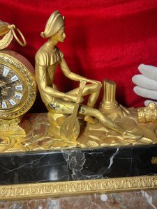 Italian Empire Figural Salon Clock With Posed Court Jester Holding Loot Circa -1950 Composition - Gilt Bronze/ Marble Listing Description A Fine Italian Gilt Bronze Timepiece Showcasing Clock Set Upon Column Mount, With Circular Bezel Face. Enamel Plaque Roman Numeral Dial With Fleur De Lys Decoration And Delicate Folate Hands. Large Ornamental Handled Urn Top Pinnacle And Unusual Sweeping Feather Plume. A Court Jester In Harlequin Costume Sits Against The Clock, Holding Loot Instrument Surrounded By Fallen Column Pillars. Figural Display Is Mounted Upon Black Marble Pediment With Gilt Bronze Frieze. Wonderful Timepiece With Rare Subject. Condition Report Signs Of Tarnishing To Gilt Work Due To Age And Usage. Dimensions In Inches Height 15" X Width 19" X Depth 8" PLEASE NOTE This Is A Very HEAVY ITEM. Due To This Heavy Nature Of This Clock, Delivery Will Be By Hand By Our Curator , Personal Collection By Buyer Or Collection By Specialist Courier Organised By The Buyer. For Further Information Or Enquiries Please Call The Curator On 07707 488566