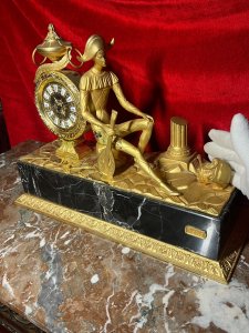 Italian Empire Figural Salon Clock With Posed Court Jester Holding Loot Circa -1950 Composition - Gilt Bronze/ Marble Listing Description A Fine Italian Gilt Bronze Timepiece Showcasing Clock Set Upon Column Mount, With Circular Bezel Face. Enamel Plaque Roman Numeral Dial With Fleur De Lys Decoration And Delicate Folate Hands. Large Ornamental Handled Urn Top Pinnacle And Unusual Sweeping Feather Plume. A Court Jester In Harlequin Costume Sits Against The Clock, Holding Loot Instrument Surrounded By Fallen Column Pillars. Figural Display Is Mounted Upon Black Marble Pediment With Gilt Bronze Frieze. Wonderful Timepiece With Rare Subject. Condition Report Signs Of Tarnishing To Gilt Work Due To Age And Usage. Dimensions In Inches Height 15" X Width 19" X Depth 8" PLEASE NOTE This Is A Very HEAVY ITEM. Due To This Heavy Nature Of This Clock, Delivery Will Be By Hand By Our Curator , Personal Collection By Buyer Or Collection By Specialist Courier Organised By The Buyer. For Further Information Or Enquiries Please Call The Curator On 07707 488566