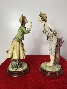 Pair Of Charming Porcelain Capodimonte Kissing Statuettes. Vintage Male And Female Pair Of Kissing Statuettes. The Lady Holds A Basket Of Oranges And Gracefully Leans In The Kiss The Male. The Male Figurine Holds A Posy Behind His Back And Leans In The Kiss His Fair Maiden. Beautiful Details, Hand Painted And Signed On The Bases. Standing On Mahogany Circular Plinths. Condition- Lovely Used Slight Damage To Finger Of Lady Otherwise Perfect Condition Dimensions In Inches Height 14 3/4” Width 5.5” Depth 5.5”