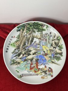 Large Porcelain Chinese Charger Plate With Ornate Celebratory Family Gathering Circa - 20th Century Vintage Composition - Porcelain Large Circular Decorative Porcelain Chinese Charger Plate With Hand Painted Scene Of A Intricate Family Celebratory Gathering Which Includes Twelve Figures Set Amongst Rocky Hillside Adorned With Oriental Trees. Some Figures Sit Playing Board Games, Reading Scriptures And Some Stand In Deep Conversation. A Young Female Carries A Laden Fruit Bowl Whilst Another Prepares Tea And Soup. Amazing Detail Of Finely Painted Faces, Hair And Hands With Wonderful Execution Of Oriental Costumes In Over Layered Fabric Kimonos And Fine Pattern In Bright Colours. Signed By Artist Front And Back. Dimensions In Inches Plate Diameter 13.5" Height From Table Top 1/2"