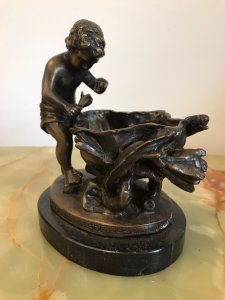 Fine Reproduction French Bronze Cherub With Flora Urn Statue. Bronze Putti In Fine Detail Holding Flower Beside A Floral Urn Decorated With Forna Vines On A Oval Black Marble Base Exceptionally Ornate With Stunning Patina Composition- Bronze Dimensions Height 6 3/4” Width 5.5” Depth 4.5” Weight 3kg Approx